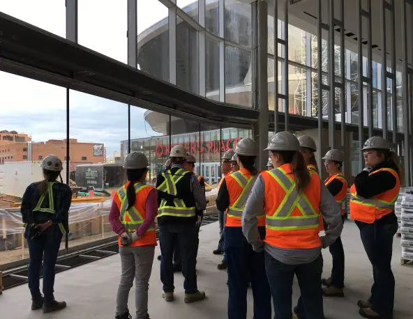 A group of construction workers wearing hard hats and high‑visibility vests stand inside a partially completed building with large glass windows, facing a construction area outside; exposed structural elements, tools, and a city street with buildings are visible in the background.
