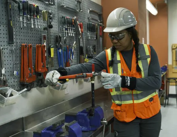 Person wearing a hard hat, safety glasses, gloves, and a high-visibility vest uses a pipe wrench to work on a metal pipe secured in a bench vise. A wall-mounted tool board with hand tools is visible in the workshop background.
