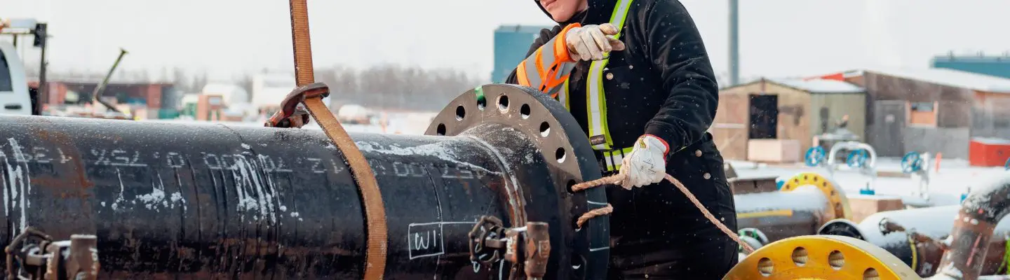 A worker wearing a hard hat, safety glasses, gloves, and a high‑visibility vest adjusts a chain or rigging on a large black industrial pipe with a bolted flange at an outdoor construction site, with snow on the ground and additional pipes and equipment in the background.