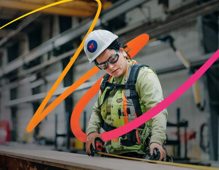 A woman in a hard hat uses a measuring tape to measure a piece of wood