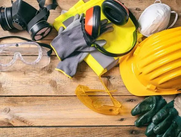 A flat lay of personal protective equipment arranged on a wooden surface, including a yellow hard hat, clear safety goggles, ear protection, a respirator mask, a white disposable mask, high‑visibility material, and several pairs of work gloves in different colors and materials.