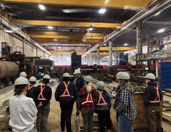 Group wearing safety helmets and high‑visibility vests gathers in a large industrial shop with metal equipment and overhead cranes.