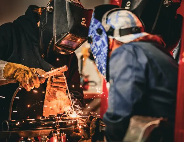Two people wearing welding helmets, protective gloves, and workwear perform a welding task on metal pipes or tubing in an indoor workshop; bright sparks and molten metal are visible at the weld point, with tools, equipment, and a warm, industrial lighting environment in the background.