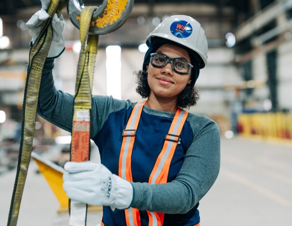 A woman wearing a hard hat, safety glasses, gloves, and a high‑visibility vest holds lifting straps inside a workshop.